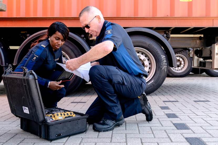 2 knielende douaniers naast een vrachtwagen, beiden in uniform met werkschoenen, donkerblauwe cargobroek en polo